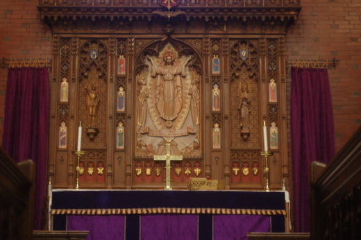 Carved wooden altar/reredos against a red brick wall, with altar table and fabrics, candlesticks, and a gold-colored metal cross