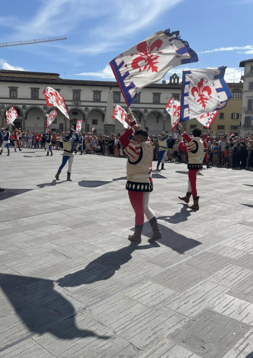 men in Mediaeval/Renaissance-style clothing wave flags in the air in an open-air city square