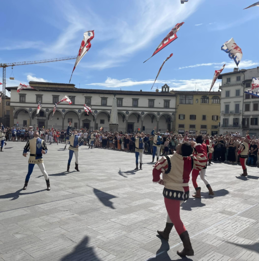 men in Mediaeval/Renaissance-style clothing toss flags into the air in an open-air city square
