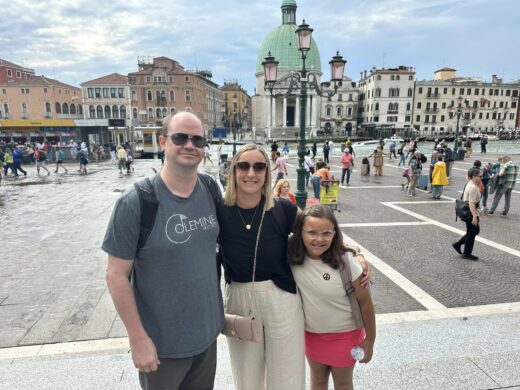 two adults and a child stand in front of a piazza or waterfront with a row of ancient buildings in the background