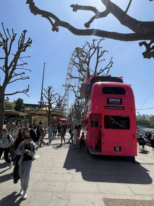 crowds on a riverside quay by a pink double-decker bus with the London Eye Ferris wheel in the near distance